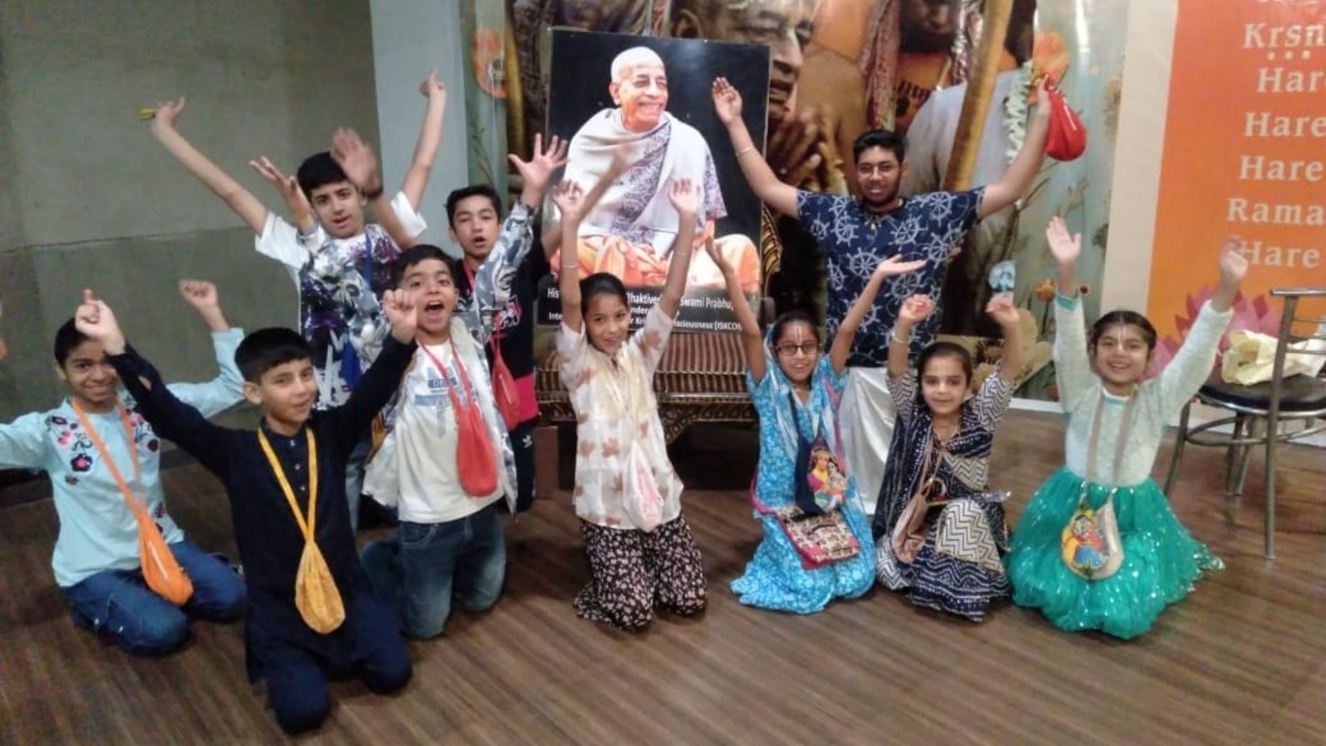 Children and mentors raising their hands in joyful Haribol celebration in front of Srila Prabhupada's portrait